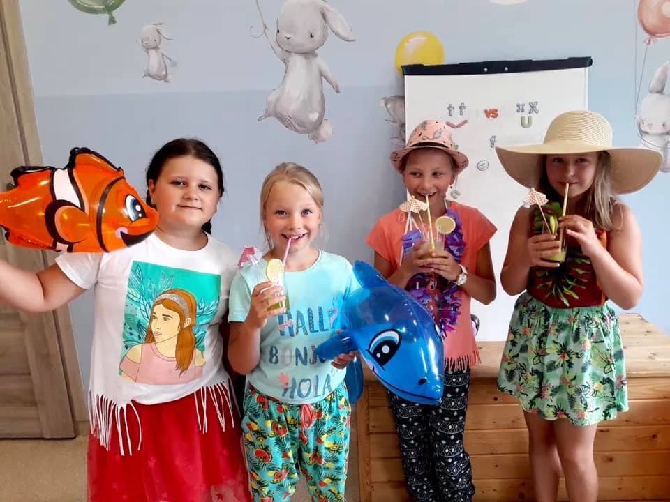 Obraz przedstawia dzieci przebrane w ubrania nawiązujące do Hawajów. Na obrazie można również zobaczyć różnego rodzaju akcesoria związane z Hawajami.