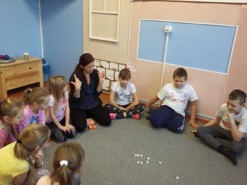 Na zdjęciu widnieje grupka dzieci siedzących na dywanie z prowadzącą zajęcia osobą. Wszyscy znajdują się w pokoju z niebieskimi ścianami.