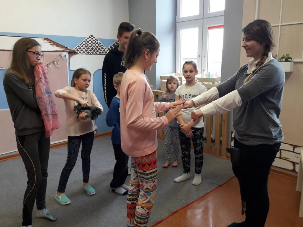 Zdjęcie przedstawia dzieci z placówki wraz z osobą prowadzącą zajęcia. Wszyscy stoją w pokoju z szarymi i białymi ścianami.