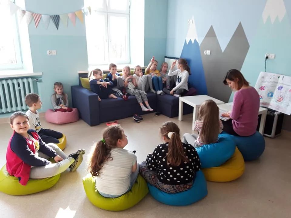Obraz przedstawia zajęcia psychoedukacyjne. Na zdjęciu możemy ujrzeć dzieci słuchające wychowawczyni,  siedzące na sofie oraz poduszkach.