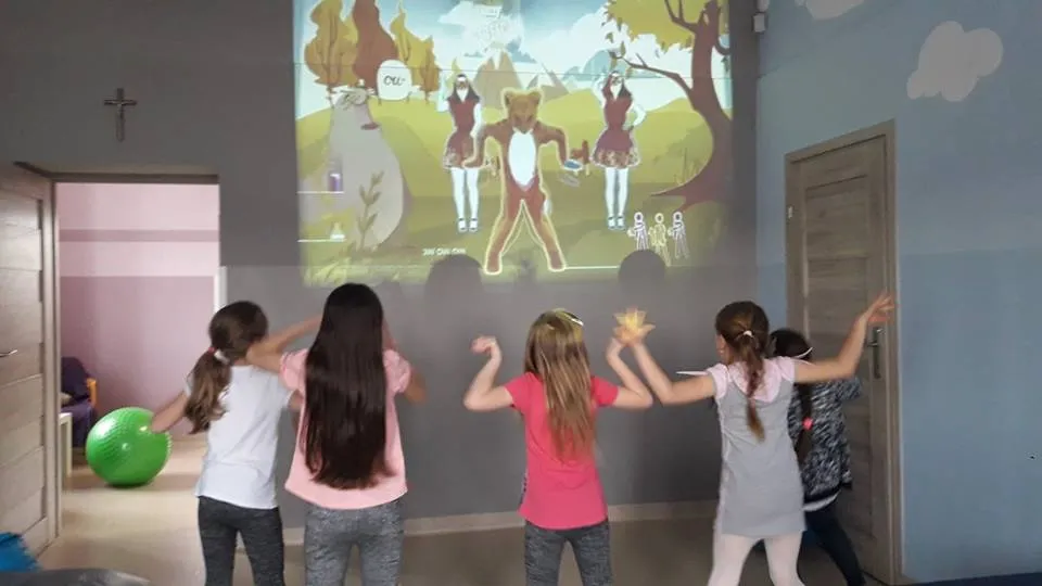 Obraz przedstawia wychowanków podczas zabaw z wykorzystaniem projektora. Na zdjęciu dzieci tańczą do filmiku wyświetlonego na ścianie przy użyciu projektora.