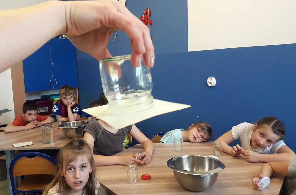 Obraz przedstawia wychowanków podczas zajęć których tematem były żywioły ziemi. Na zdjęciu możemy ujrzeć zaciekawione dzieci, aktualnym eksperymentem. Wychowankowie siedzą przy stołach, na których są przygotowane przedmioty do doświadczeń. 