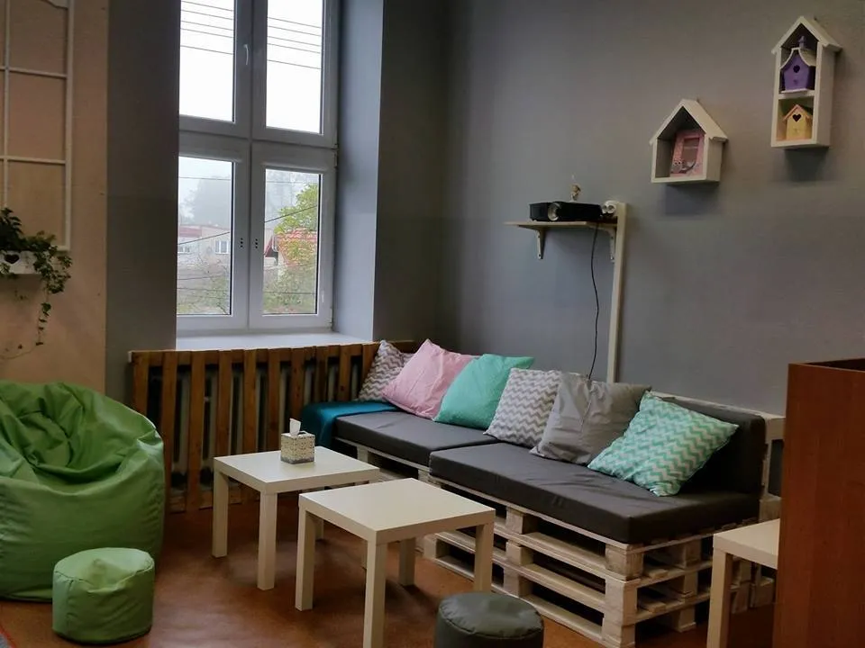 Na zdjęciu widnieje odnowiony pokój w SPWD w Radłowie. Pomieszczenie zawiera: zielone siedzisko dla dzieci, 2 stoliki, kanapa z palet, kilka poduszek na niej, ściany są koloru szarego z powieszonymi półkami w kolorze białym, dodatkowo poczwórne okno.