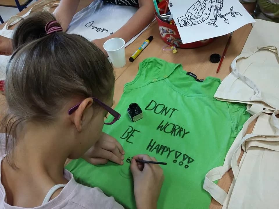 Obraz przedstawia dziewczynkę malującą na zielonej koszulce położonej na stole napis "Dont worry, be happy!"