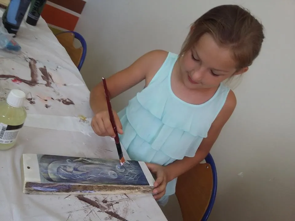 Zdjęcie przedstawia dziewczynkę malującą coś na drewnie. Stół jest przykryty białym obrusem.