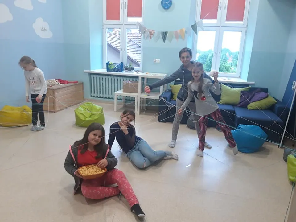 Obraz przedstawia wychowanków SPWD podczas zabaw, gdy na zewnątrz pada deszcz. Na zdjęciu możemy ujrzeć dzieci bawiące się na torze przeszkód wykonanym z włóczek.