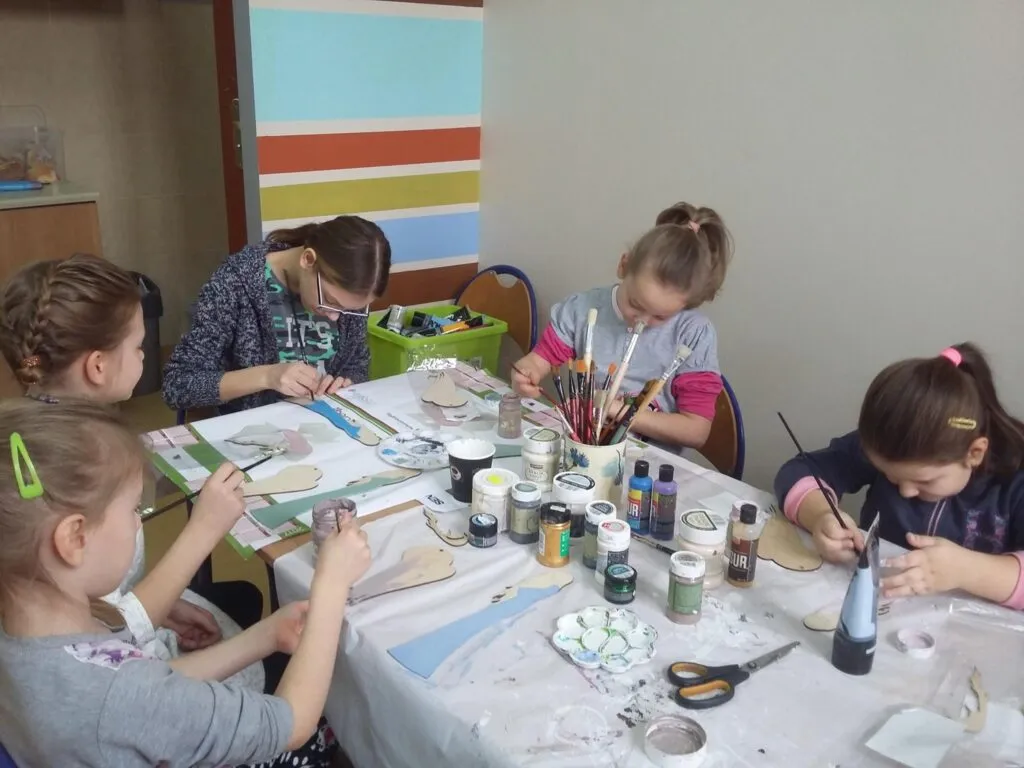 Zdjęcie przedstawia dzieci z placówki przy stole z białym nakryciem tworzących kolejne rękodzieła.