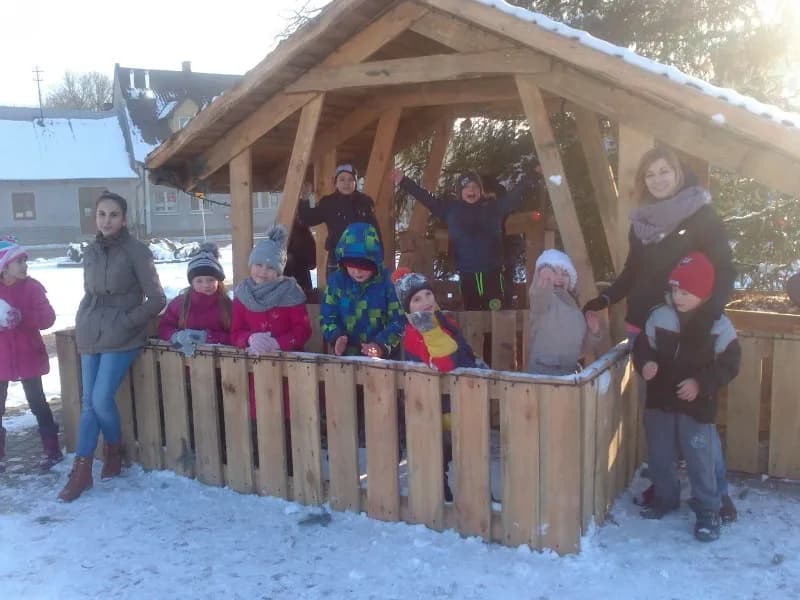 Obraz przedstawia dzieci z placówki cieszące się śniegiem. Pozują do zdjęcia w niewielkim drewnianym budynku.