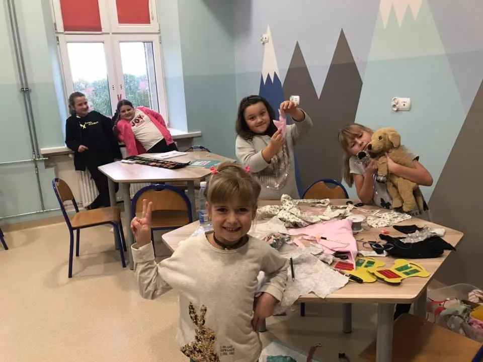 Obraz przedstawia dzieci w placówce, które tętnią radością wykonując różnego rodzaju prace plastyczne.