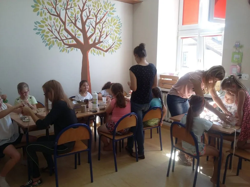 Zdjęcie przedstawia dzieci siedzące przy stolikach w sali z namalowanym drzewem