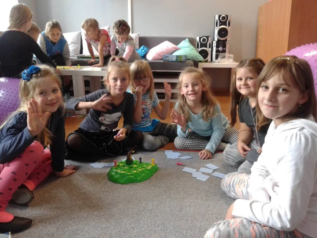 Na zdjęciu widnieje grupka dzieci siedzących na dywanie oraz grających w grę planszową. W oddali widać drugą grupkę dzieci, które również grają w inną grę.