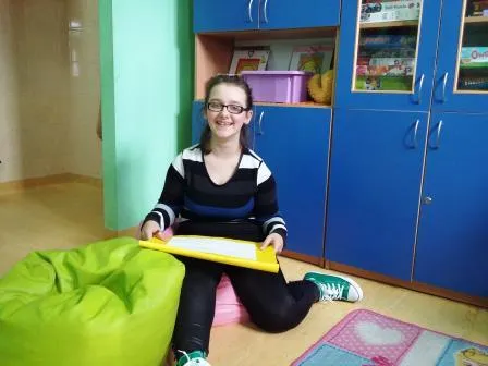 Zdjęcie przedstawia dziewczynkę siedzącą na tle niebieskich szafek, która trzyma w rękach podkładkę a na niej kartkę papieru.