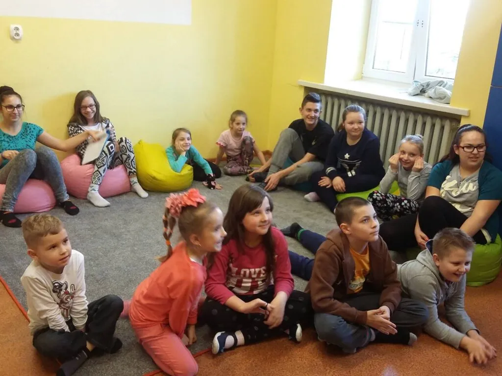 Zdjęcie przedstawia dzieci z placówki siedzące na dywanie w pomieszczeniu z żółtymi ścianami podczas zajęć.